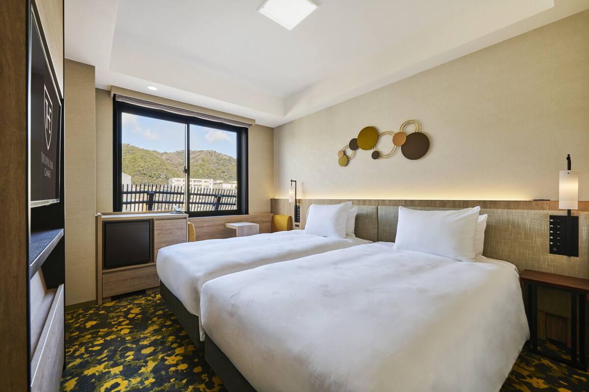 Habitación de hotel con dos camas, un escritorio, una silla, un ventanal con vistas a la montaña y un adorno en la pared.