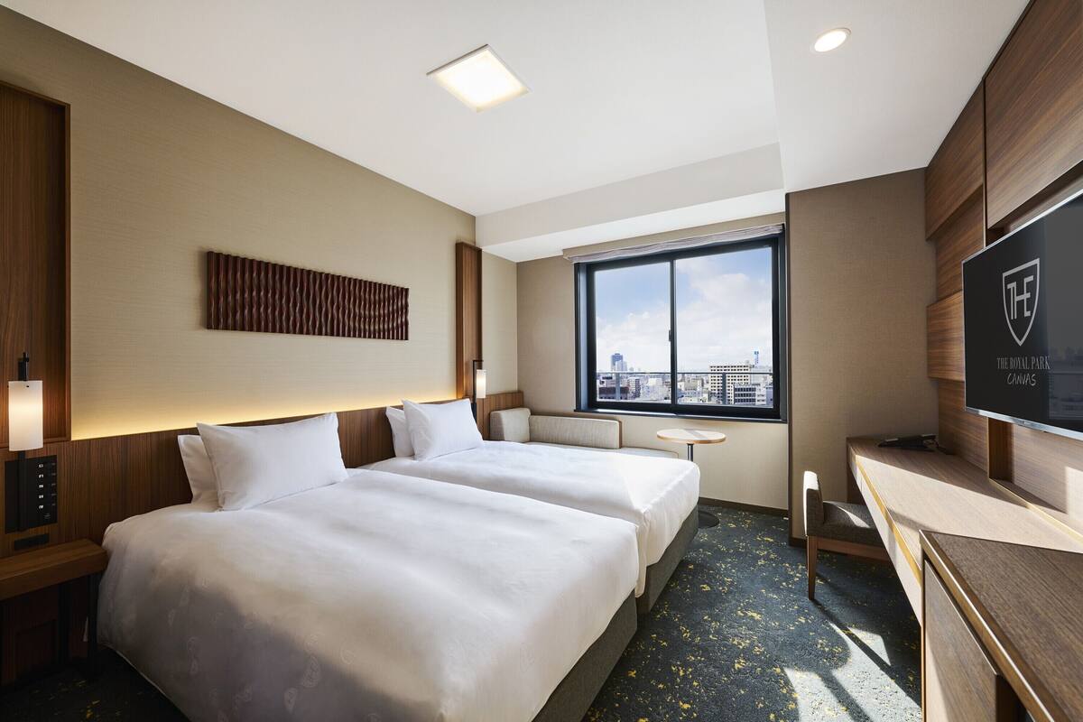 Habitación de hotel con dos camas, un escritorio, televisión, ventana con vista a la ciudad y una obra de arte en la pared.