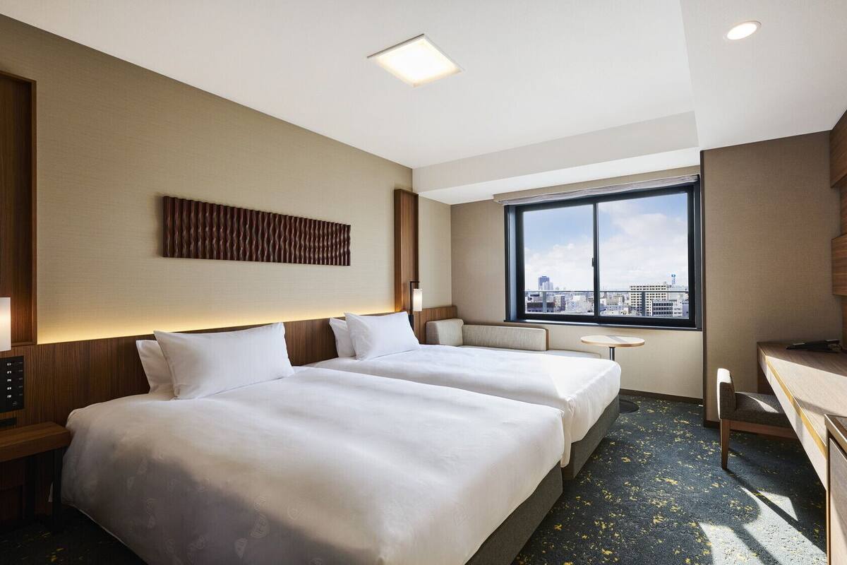 Habitación de hotel con dos camas, un escritorio, un televisor y un ventanal con vistas a la ciudad.