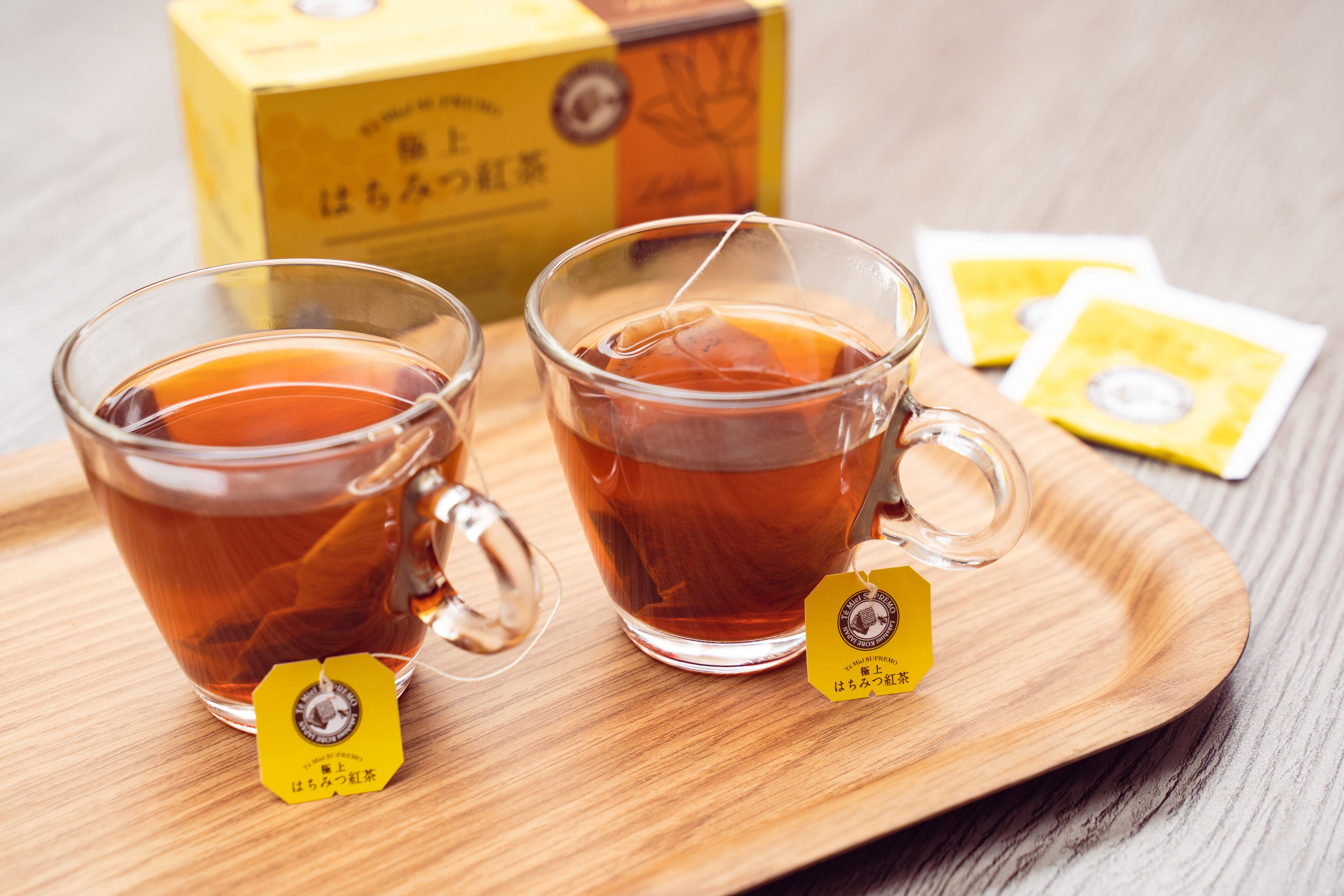 Dos tazas de té de vidrio sobre una bandeja de madera, con bolsitas de té y una caja al fondo.
