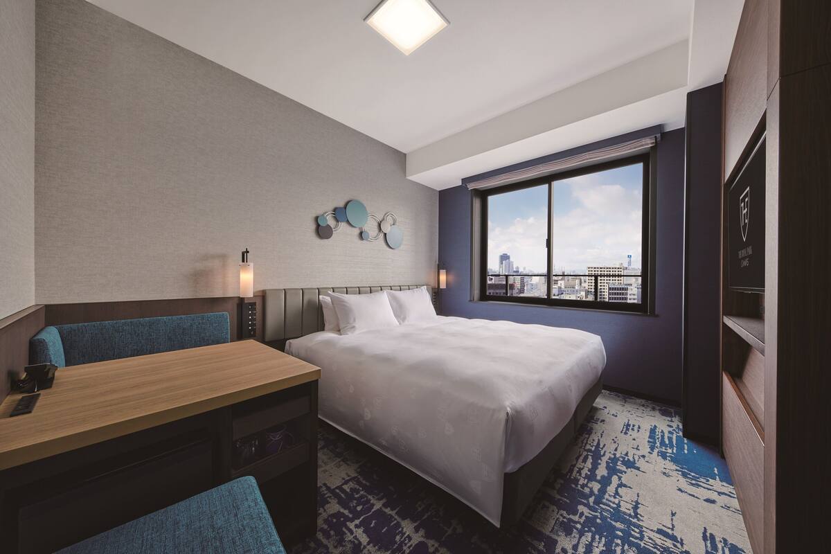 Habitación de hotel con cama, escritorio, silla, ventana grande con vista a la ciudad y una obra de arte colgada en la pared.