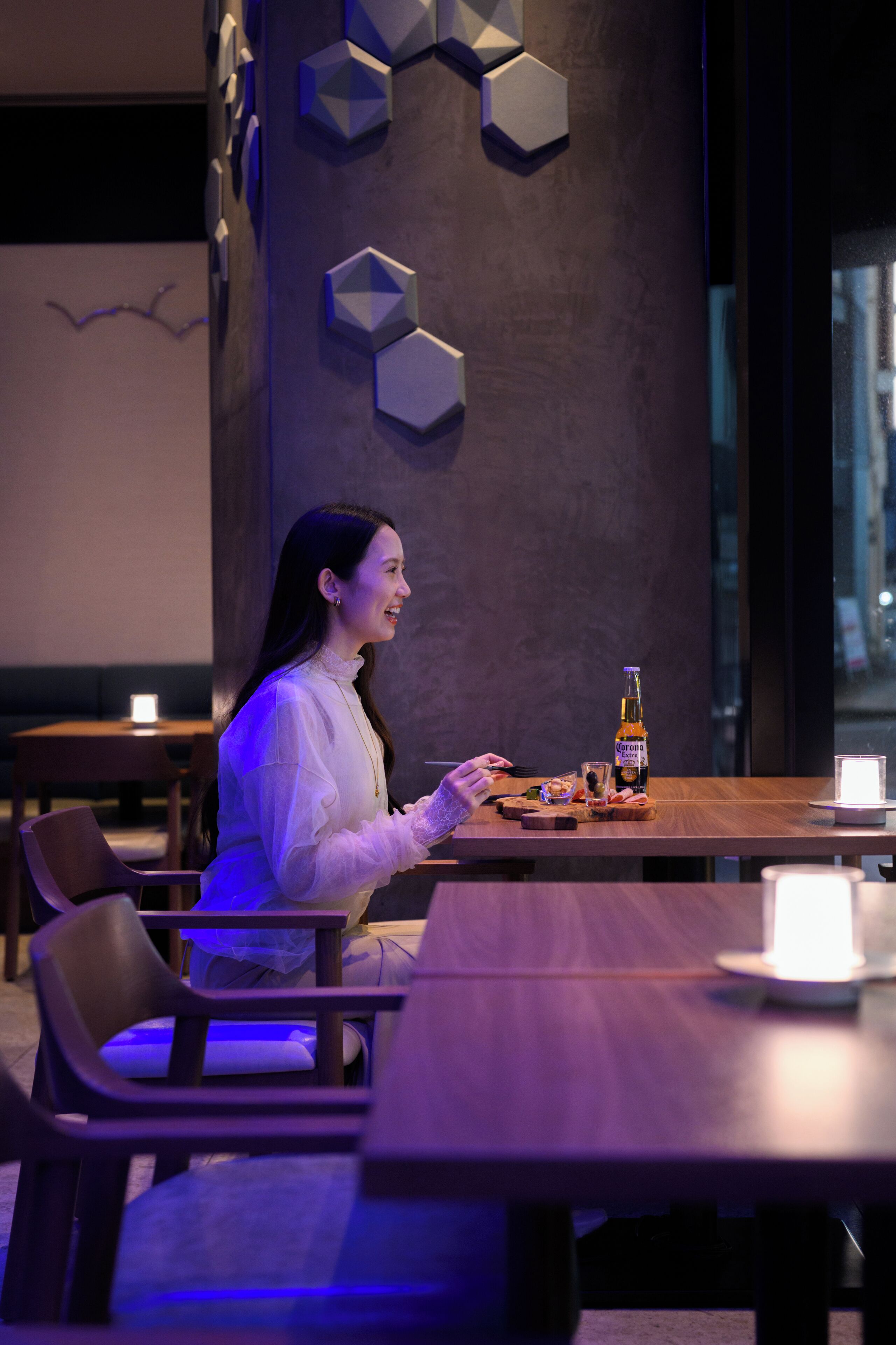 Une femme est assise à une table dans une pièce faiblement éclairée, devant une bouteille de bière Corona, un verre et une bougie.
