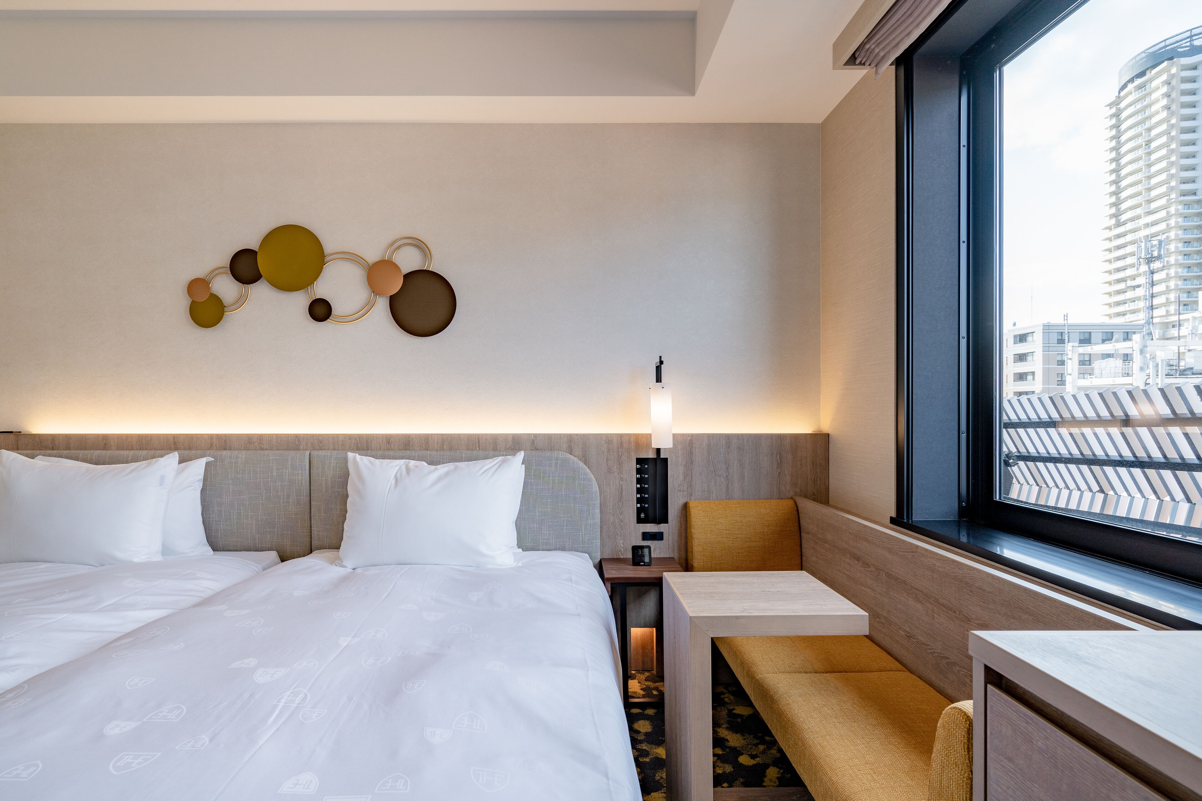 A hotel room with a large bed, a desk, a chair, a wall-mounted lamp, and a window with a view of buildings.