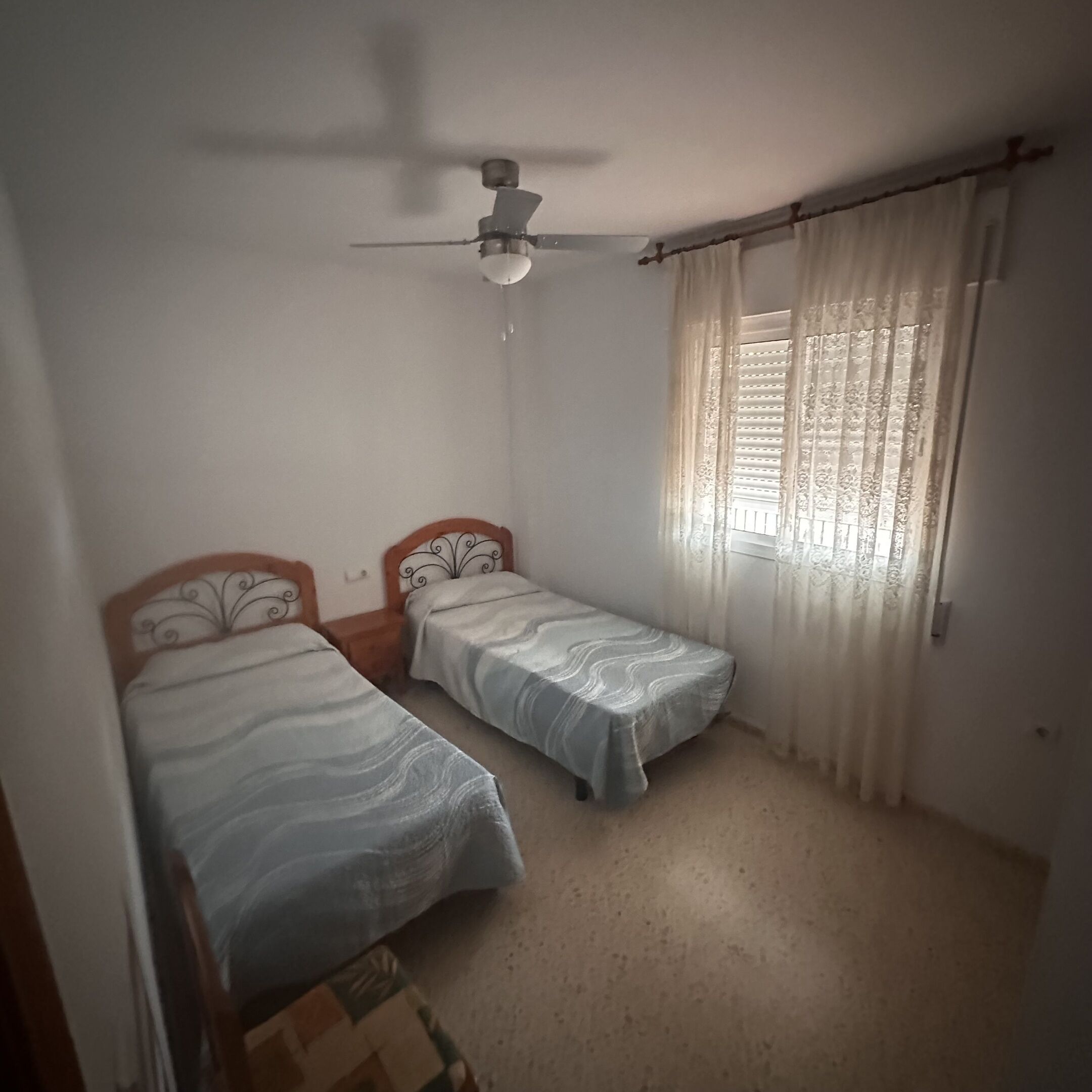 Una habitación pequeña y sencilla con dos camas individuales, un ventilador de techo y una ventana con cortinas.