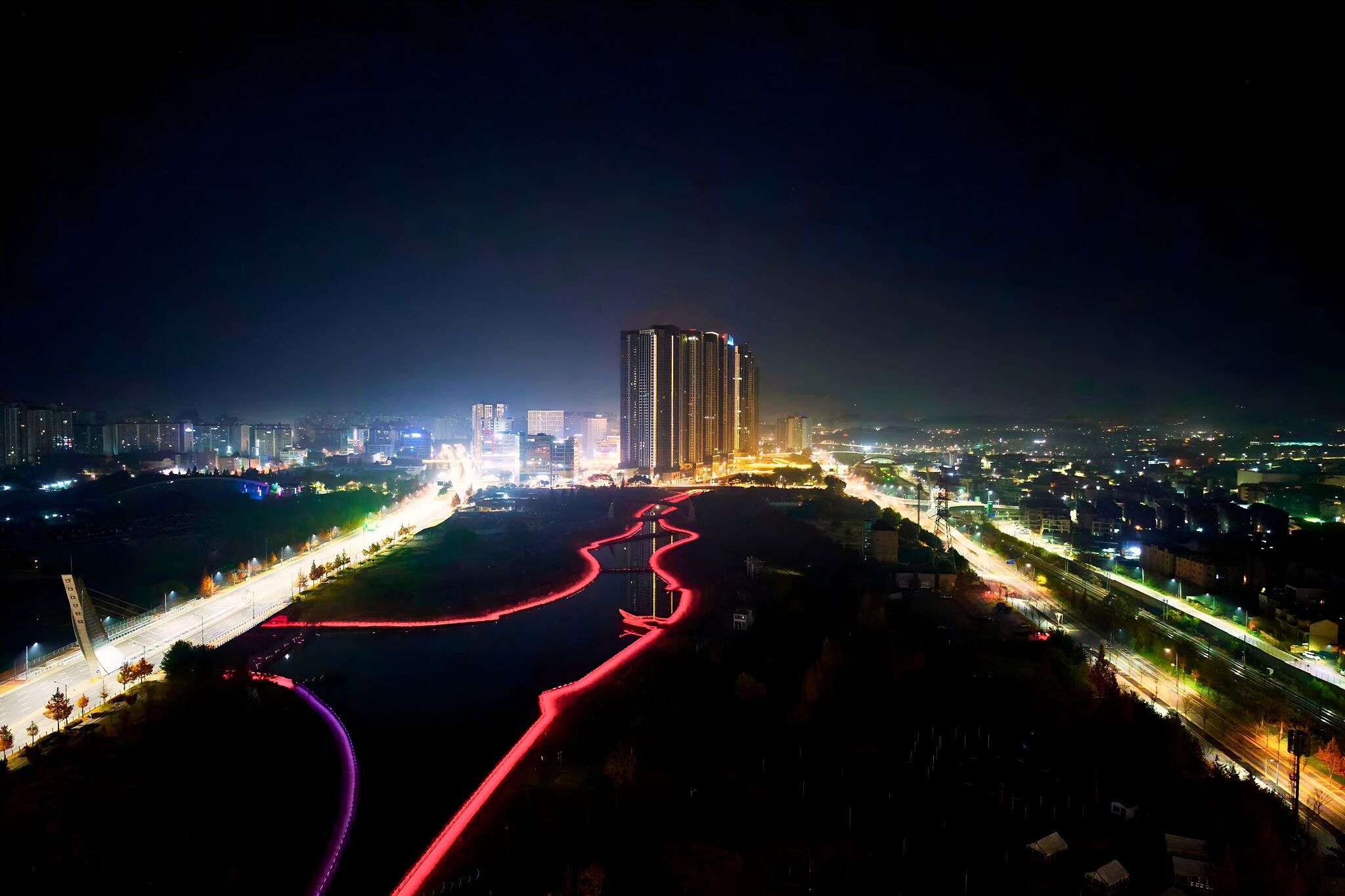 Un paisaje urbano nocturno con un rascacielos destacado, calles iluminadas y un río con estelas de luz.