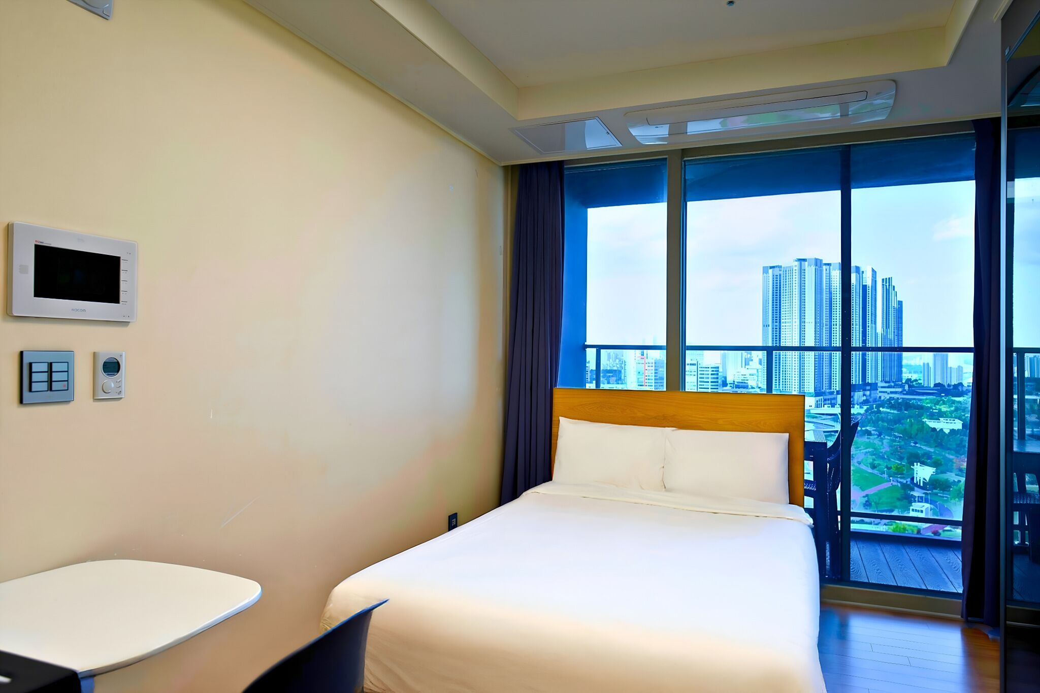 Habitación de hotel con cama, una mesita, panel de control en la pared y un ventanal con vistas a la ciudad.