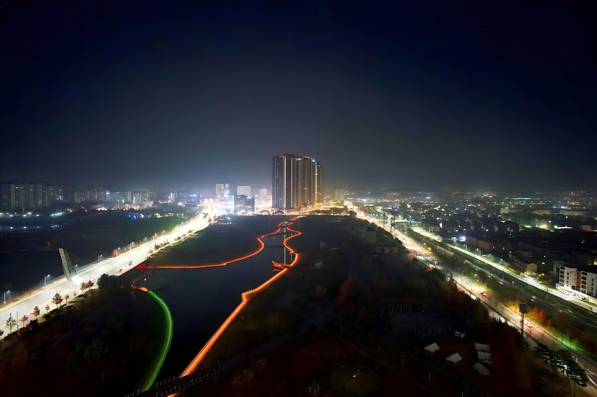 An aerial view of a cityscape at night with illuminated roads, a river, and high-rise buildings.