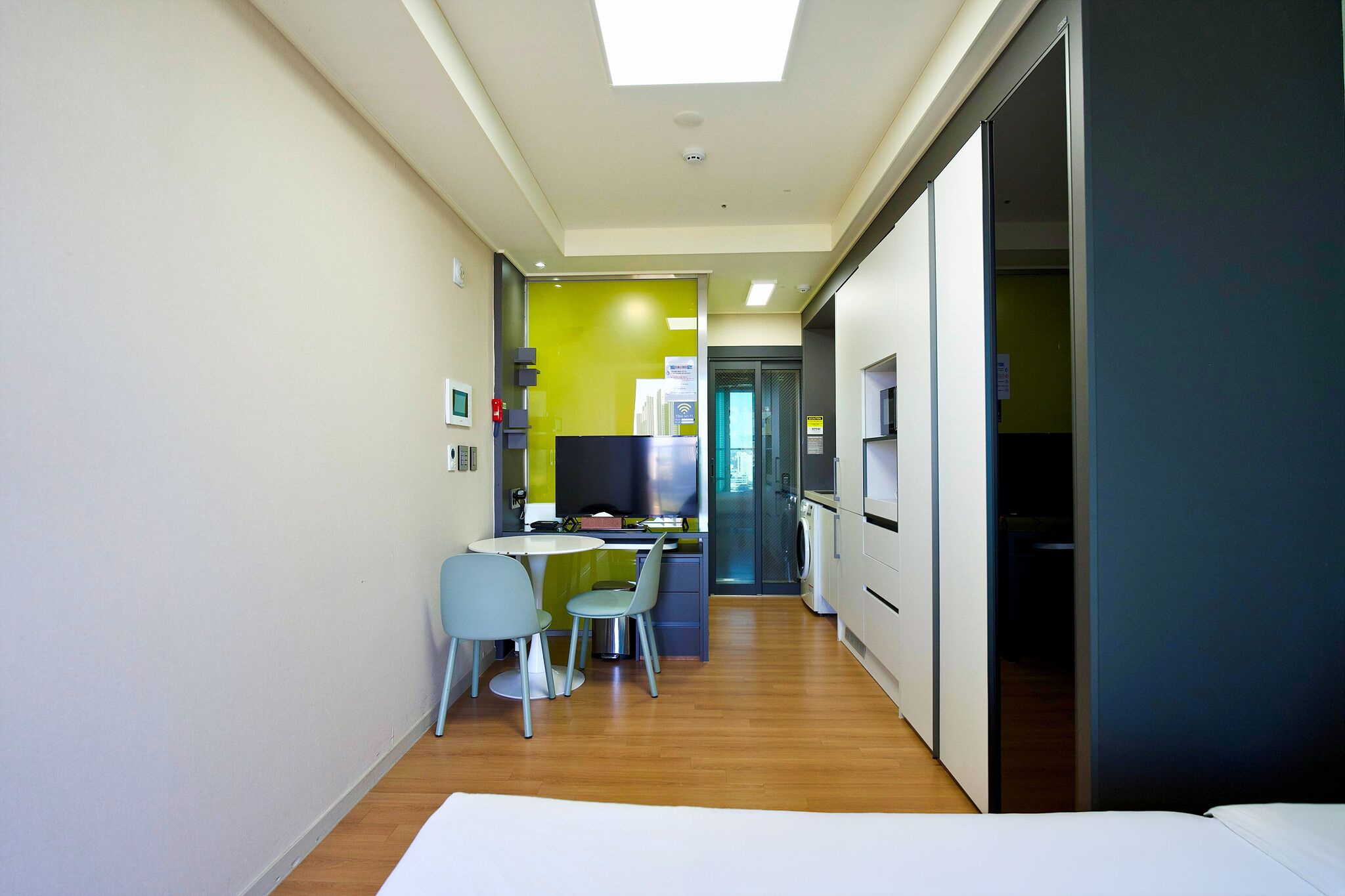 A modern hotel room with a bed, a small dining area with a table and chairs, a kitchenette, and a bathroom visible through an open door.