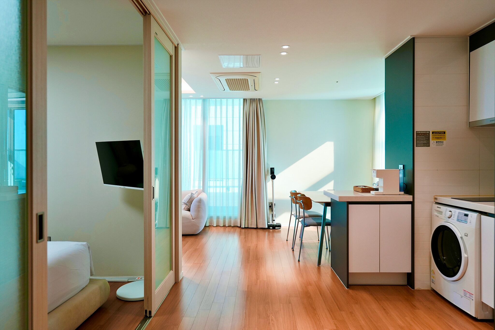 A modern hotel room with a bed, a TV, a dining area with chairs, a washing machine, and a large window with curtains.