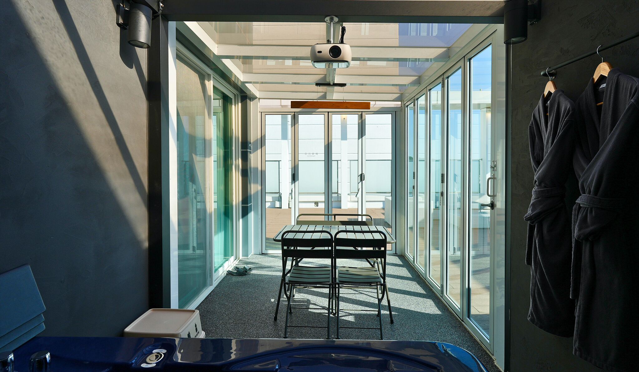 A modern, well-lit room with a glass door leading to an outdoor area, a small table with chairs, and a robe rack.