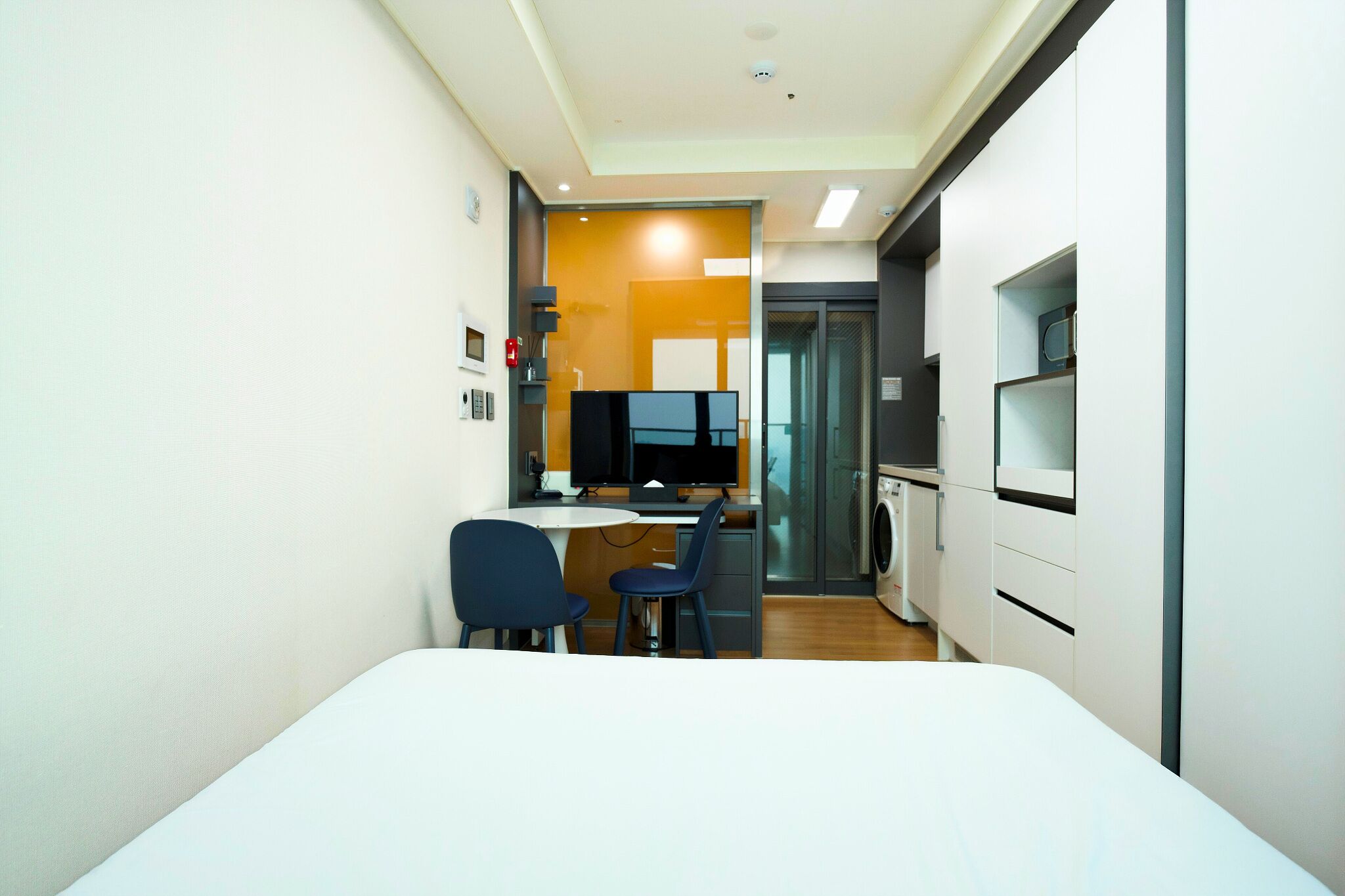 A compact hotel room with a bed, a desk, two chairs, a TV, a washing machine, and a kitchenette.