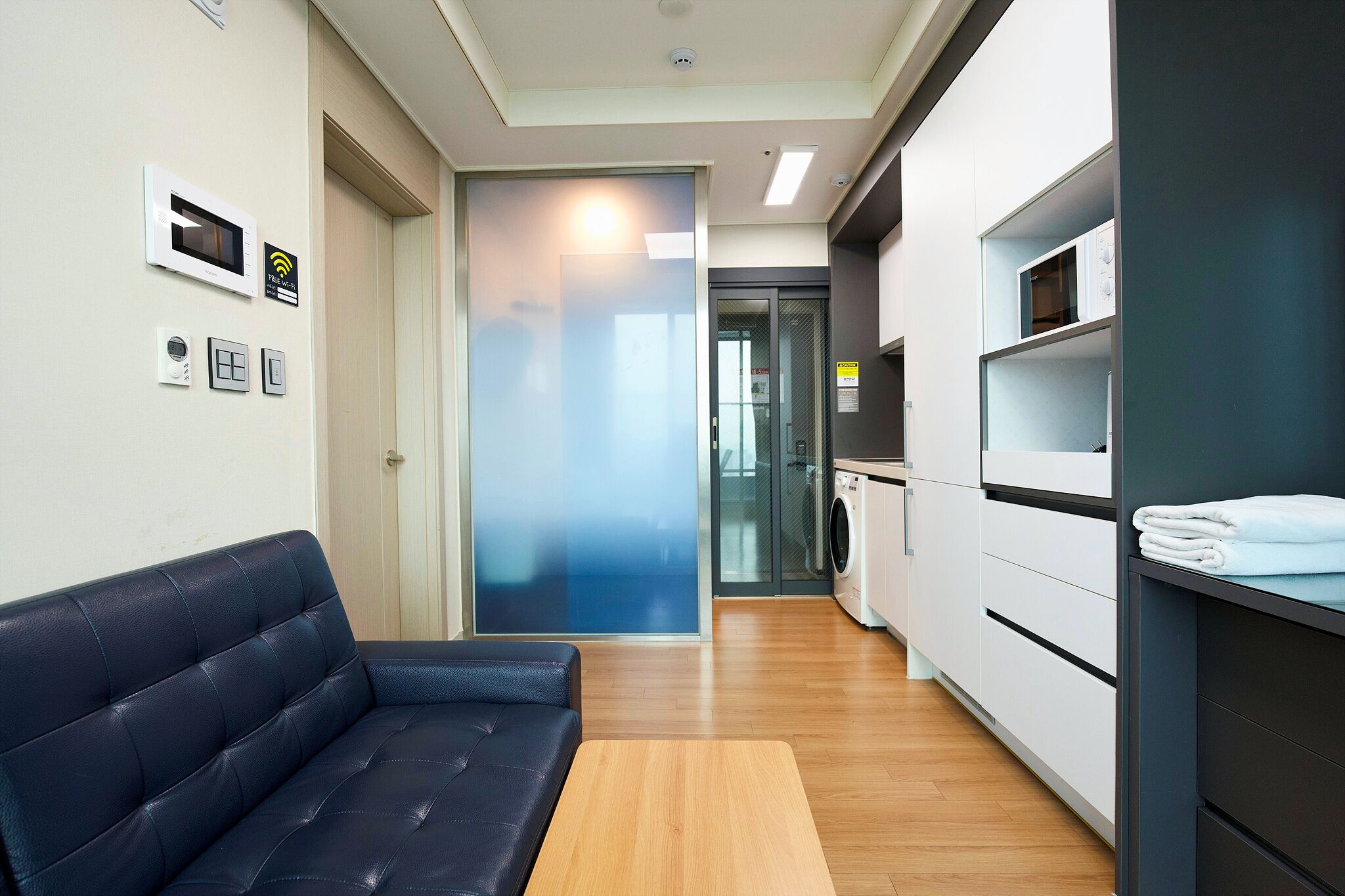 Habitació d'hotel moderna amb un sofà, una taula de fusta, una zona de cuina i una porta de vidre que dona a una altra habitació.