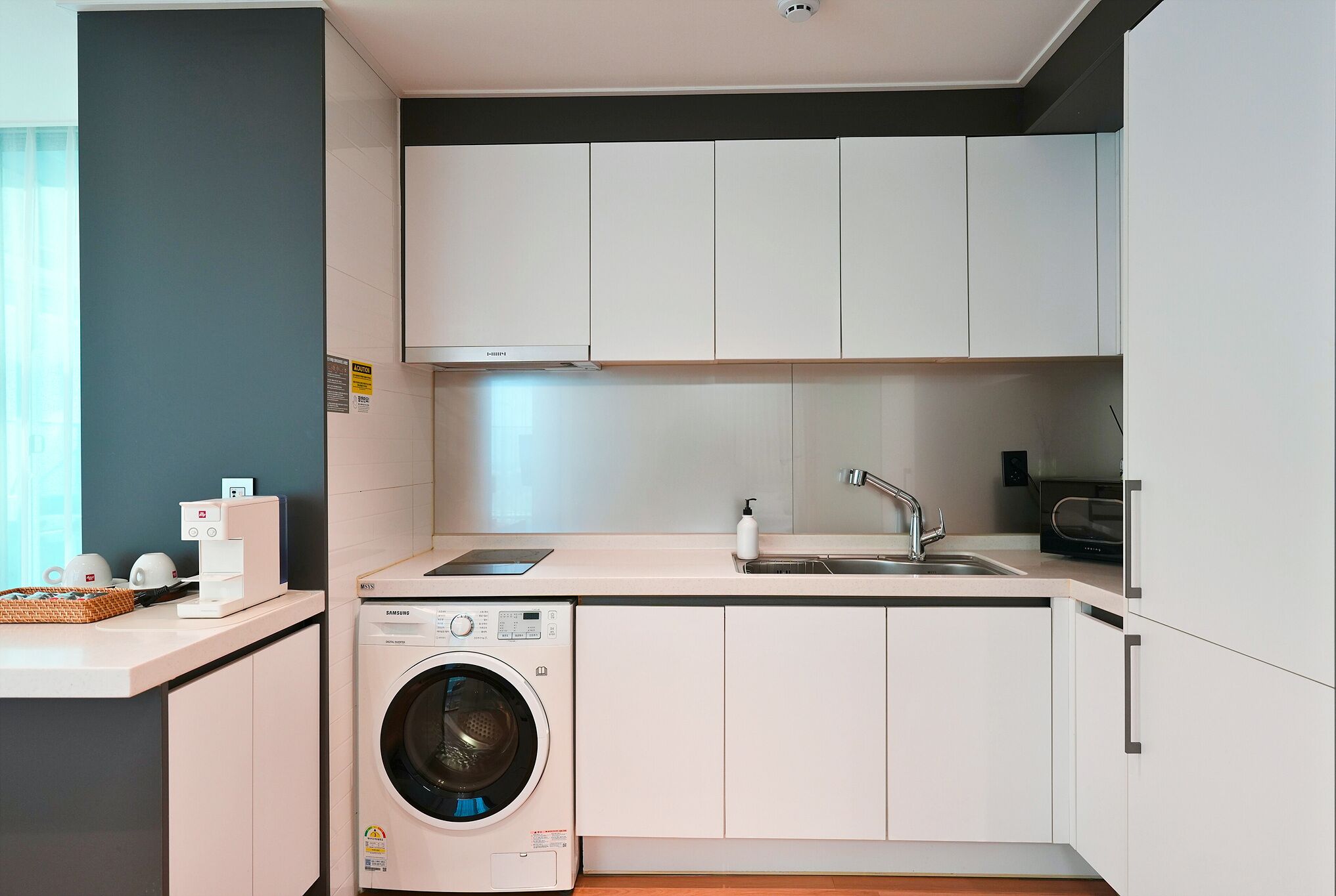 Una cocina compacta con armarios blancos, lavadora integrada, microondas y un lavamanos.