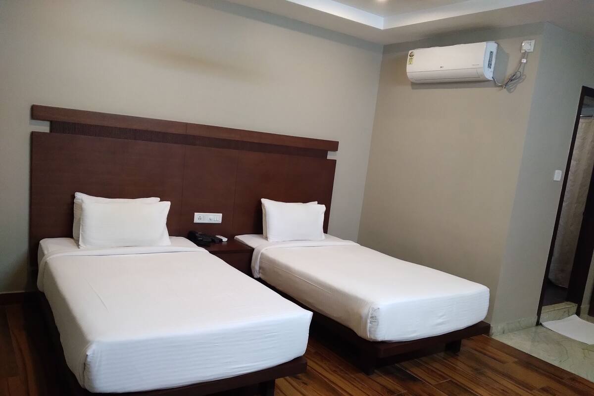 A hotel room with two single beds, a wooden headboard, a nightstand, a wall-mounted air conditioner, and a wooden floor.