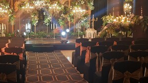 An indoor venue with rows of chairs, a stage with a backdrop of flowers and greenery, and chandeliers. - Hotel MD's Continental (East Jorhat)