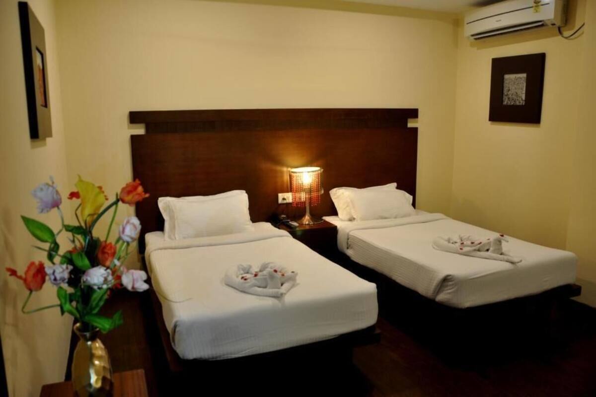 A hotel room with two single beds, a nightstand with a lamp, a vase of flowers, and an air conditioning unit.