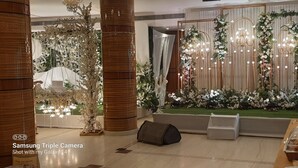 A banquet hall with a stage decorated with floral arrangements and chandeliers, a buffet area with food containers, and a large pillar. - Hotel MD's Continental (East Jorhat)