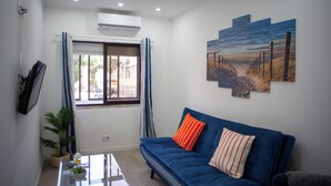 A living room with a blue sofa, a flat-screen TV on the wall, a window with curtains, and a painting of a beach scene. - Just4u Apartment - Near Lisbon & Beach - With AC (Almada)