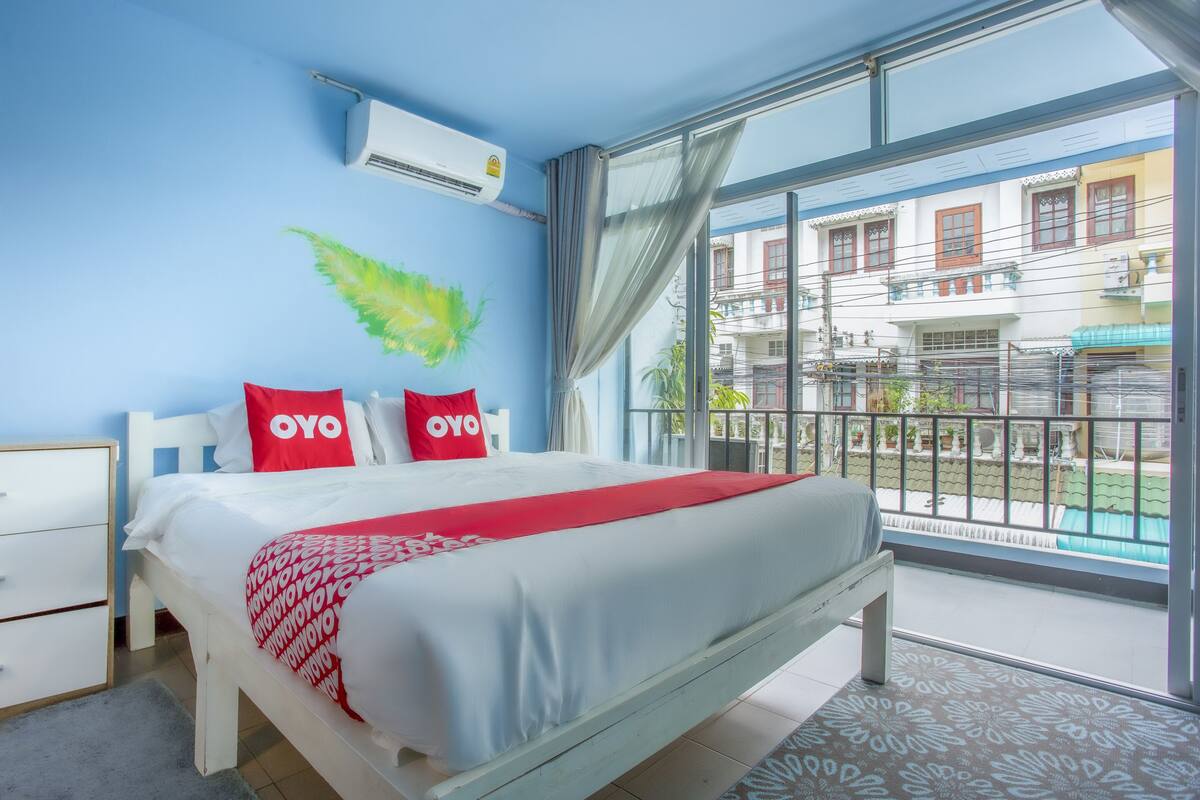 Une chambre à coucher comprenant un lit, une table de chevet, une fenêtre donnant sur des immeubles et un appareil de climatisation.