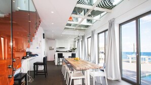 Een moderne keuken met een houten aanrecht en witte stoelen, een eethoek met een lange tafel, en grote ramen met uitzicht op een balkon en de zee.