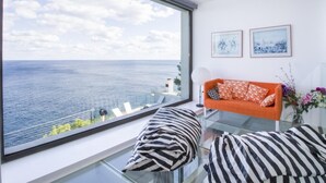 Een moderne woonkamer met een groot raam met uitzicht op zee, een glazen tafel en een bank met gestreepte kussens.