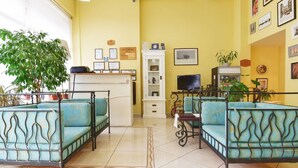 Een hotellobby met gele wanden, een receptiebalie, een zithoek met blauwe gestreepte banken, een grote potplant en ingelijste foto’s aan de wanden.