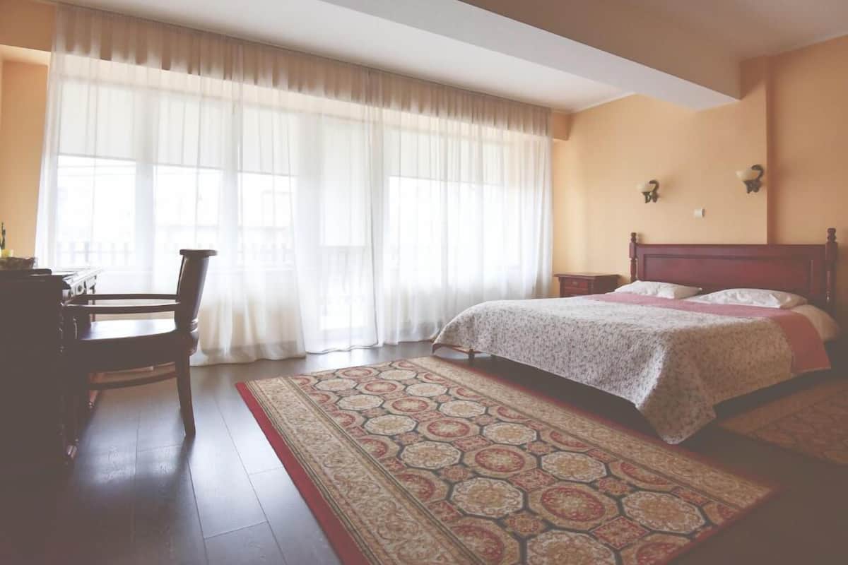 Quarto com cama, cadeira, escrivaninha e janela com cortinas leves.