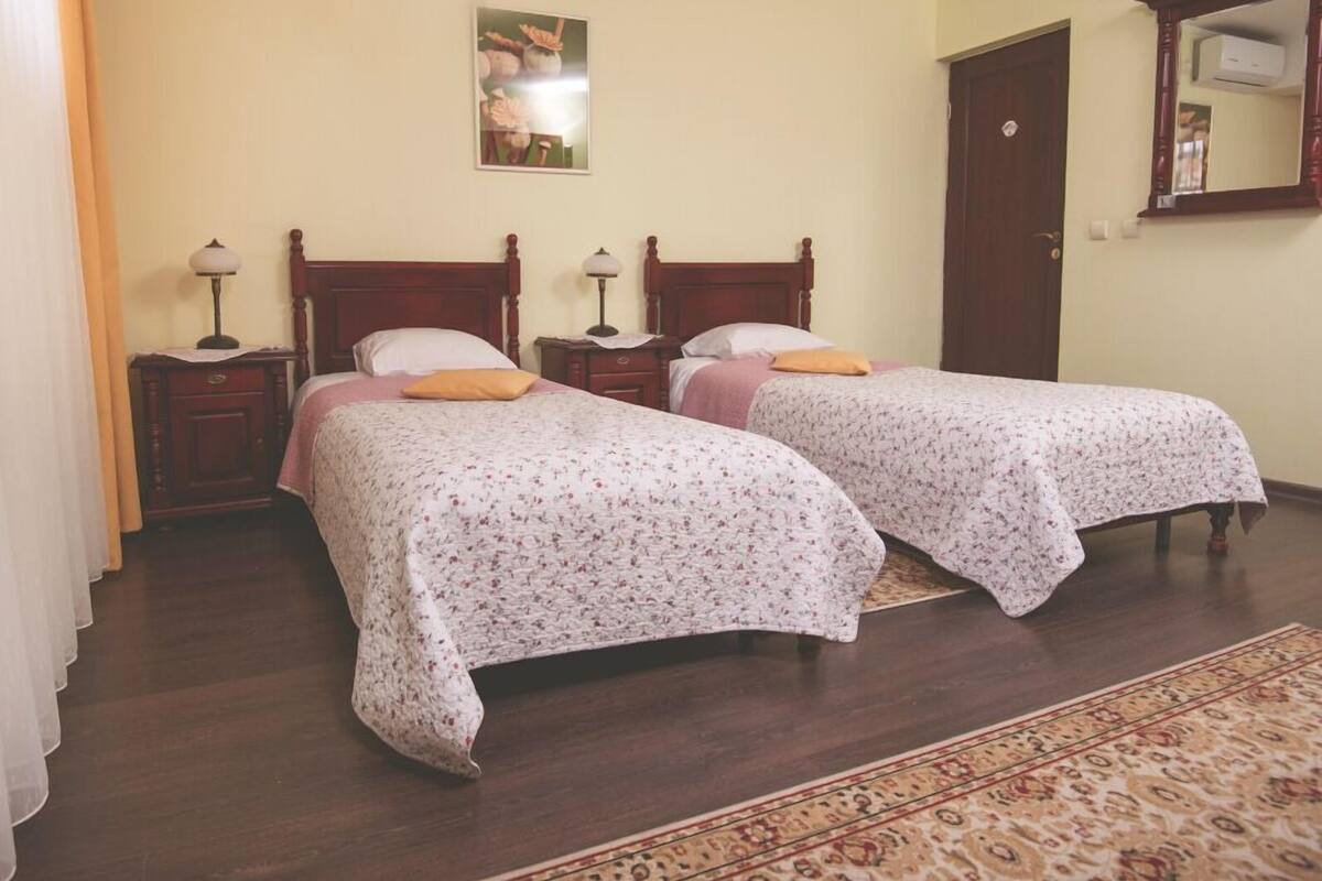 Quarto com duas camas de madeira, cada uma com colcha floral e uma almofada. Em cada lado das camas, há uma mesa de cabeceira com uma luminária. Um espelho grande está pendurado na parede acima da porta, e um pequeno porta-retrato é visível na parede.