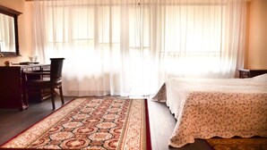 Een slaapkamer met een bed, een bureau, een stoel en een groot raam met dunne gordijnen.