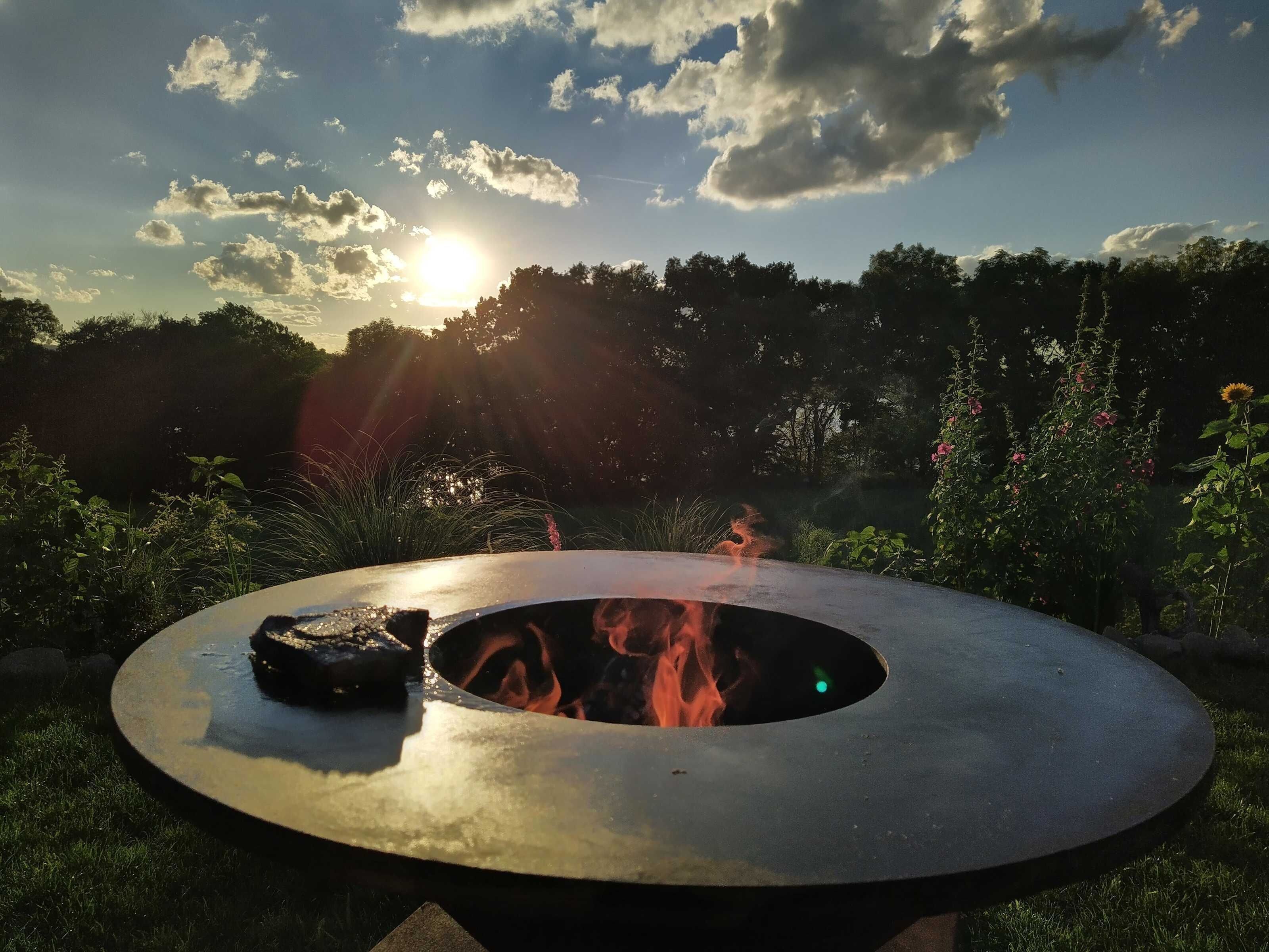 Un foyer extérieur circulaire, avec des flammes, entouré de verdure et d’arbres.