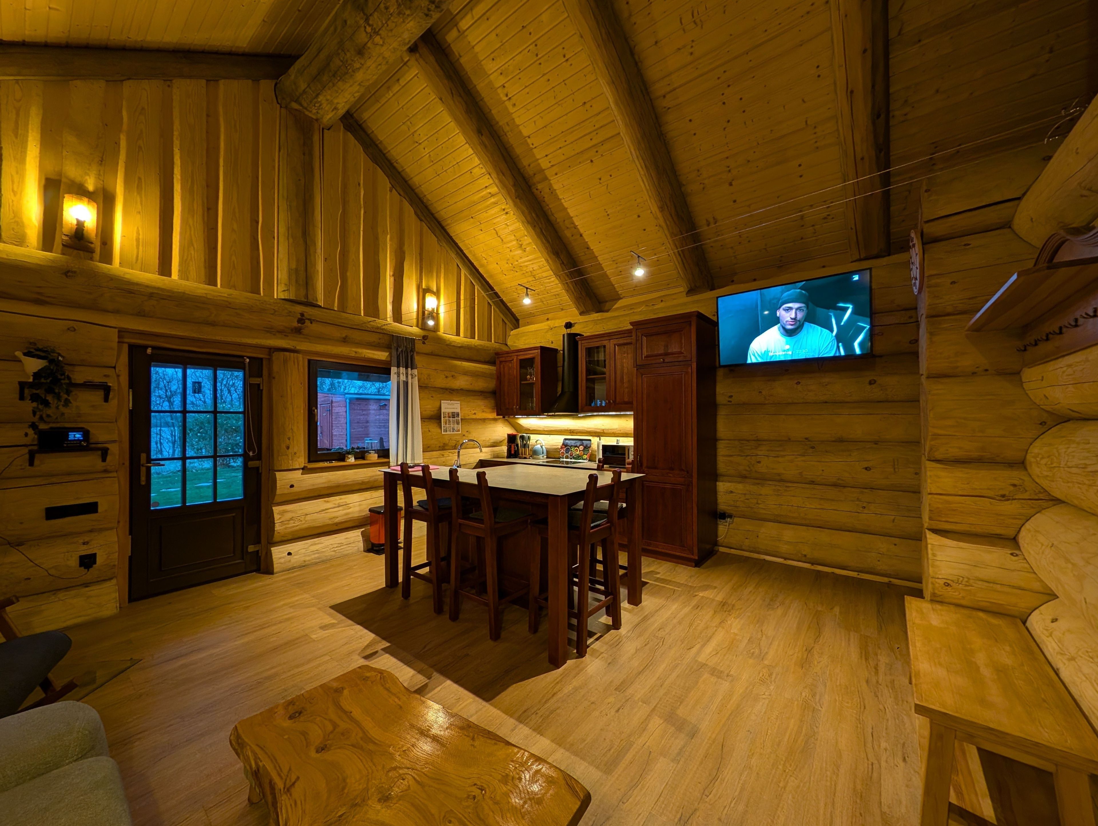 L’intérieur d’une cabane en rondins comprend un coin repas, des murs en bois, un téléviseur fixé au mur et une fenêtre avec des rideaux.