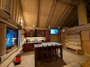 Interior de una cabaña de troncos con cocina, que incluye armarios de madera, estufa, microondas y una mesa de comedor con sillas.
