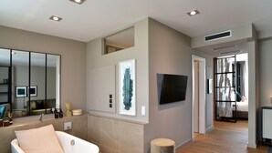Une salle de bain moderne avec une baignoire, un miroir et des toilettes. On y trouve un petit tabouret rond et une œuvre d’art encadrée accrochée au mur.
