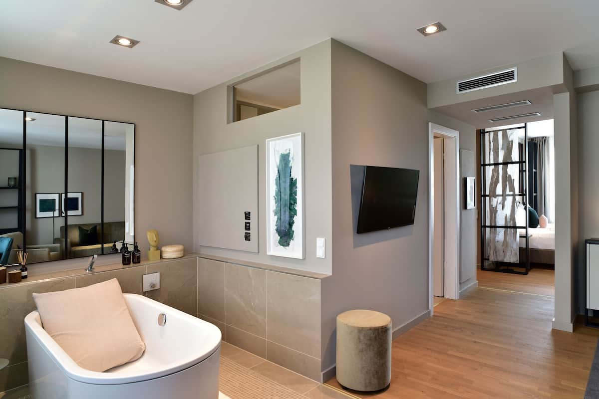Une salle de bain moderne avec une baignoire, un miroir et des toilettes. On y trouve un petit tabouret rond et une œuvre d’art encadrée accrochée au mur.