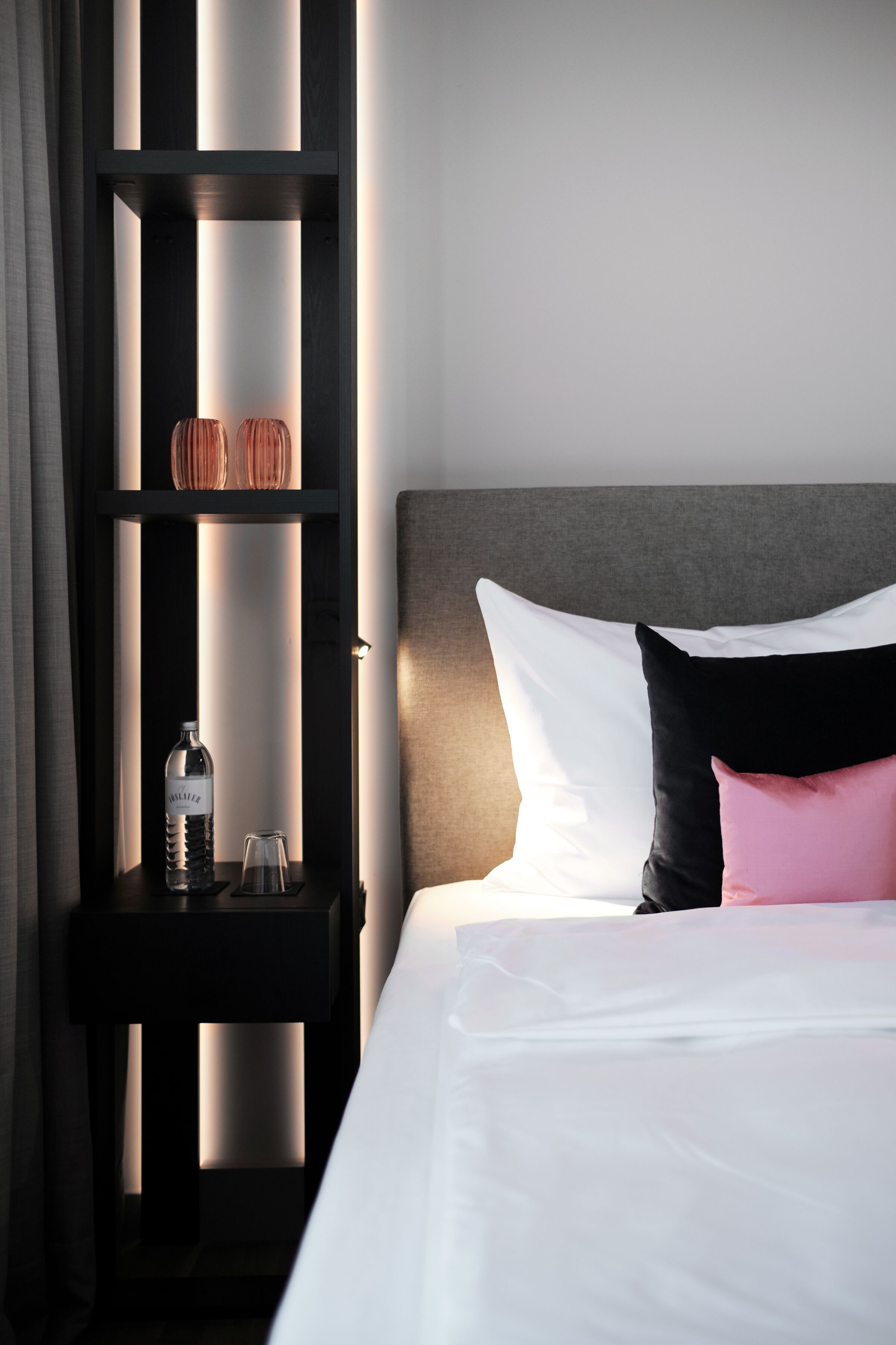 A neatly made bed with white and pink pillows, a dark nightstand with a bottle and glass, and a shelf with decorative items.