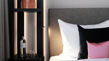 A neatly made bed with white and pink pillows, a dark nightstand with a bottle and glass, and a shelf with decorative items.