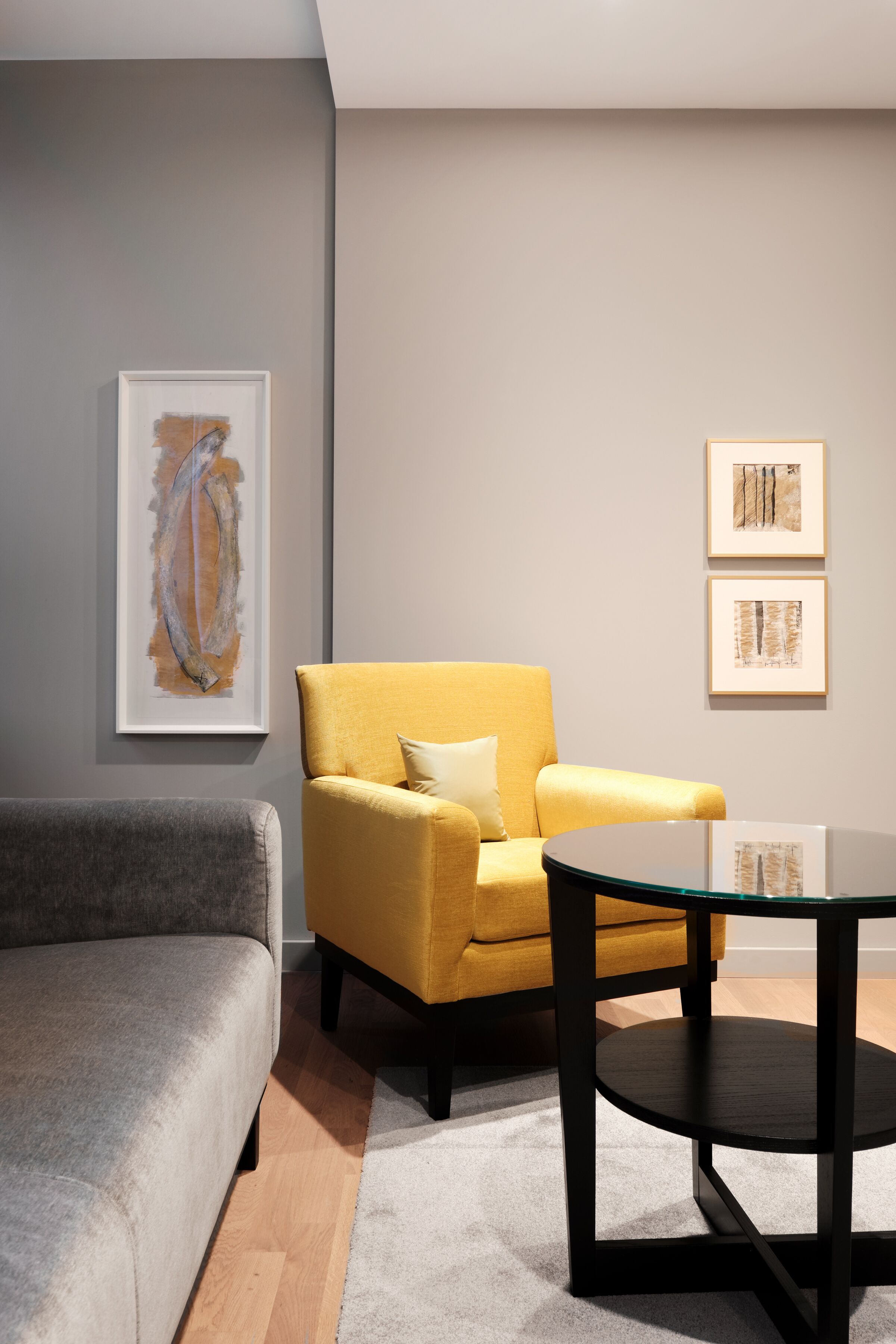 Ein modernes Wohnzimmer mit einem grauen Sofa, einem gelben Sessel und einem Couchtisch mit Glastischplatte. An den Wänden befinden sich gerahmte Kunstwerke.