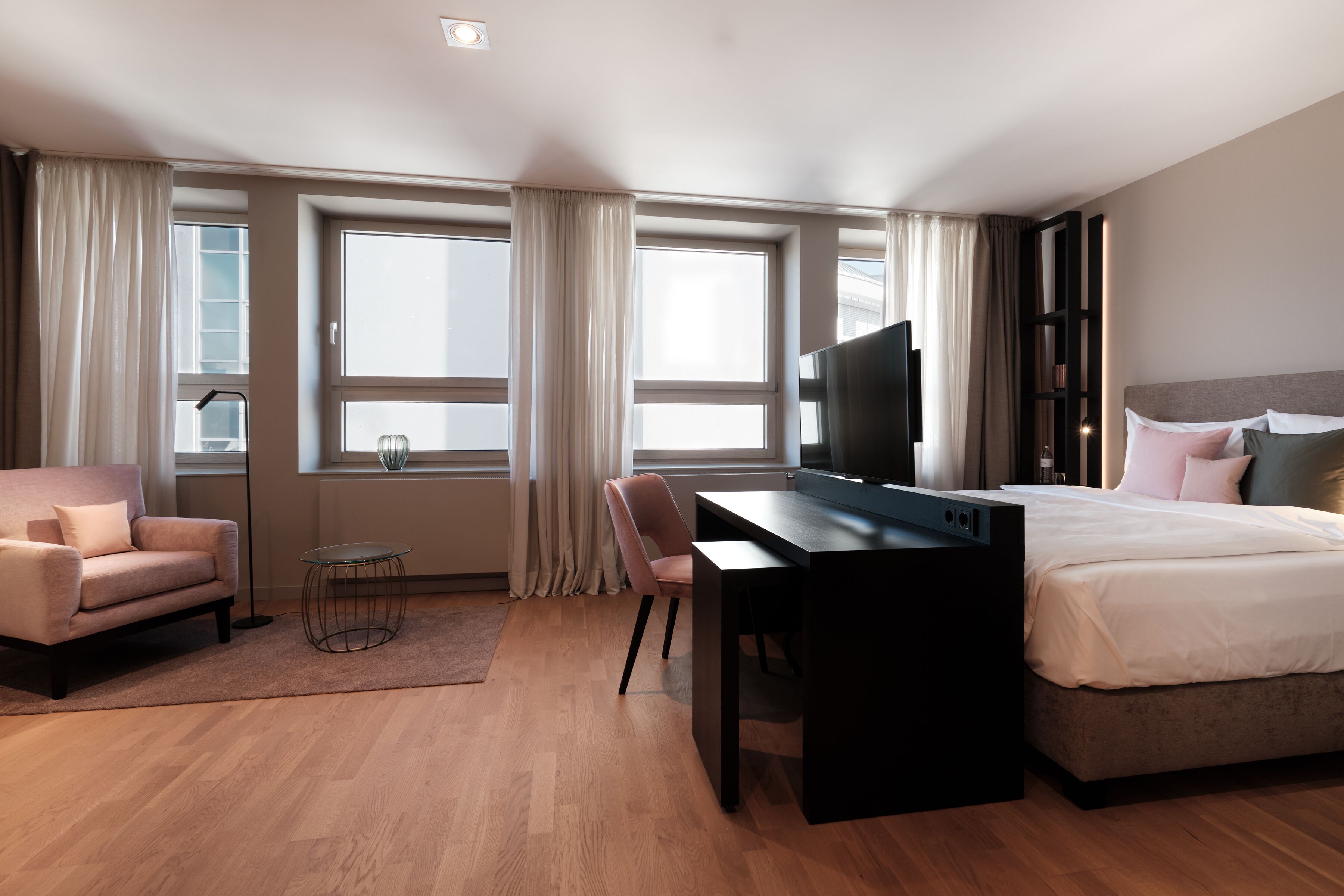 A modern hotel room with a bed, a desk, a chair, a TV, and large windows with curtains.