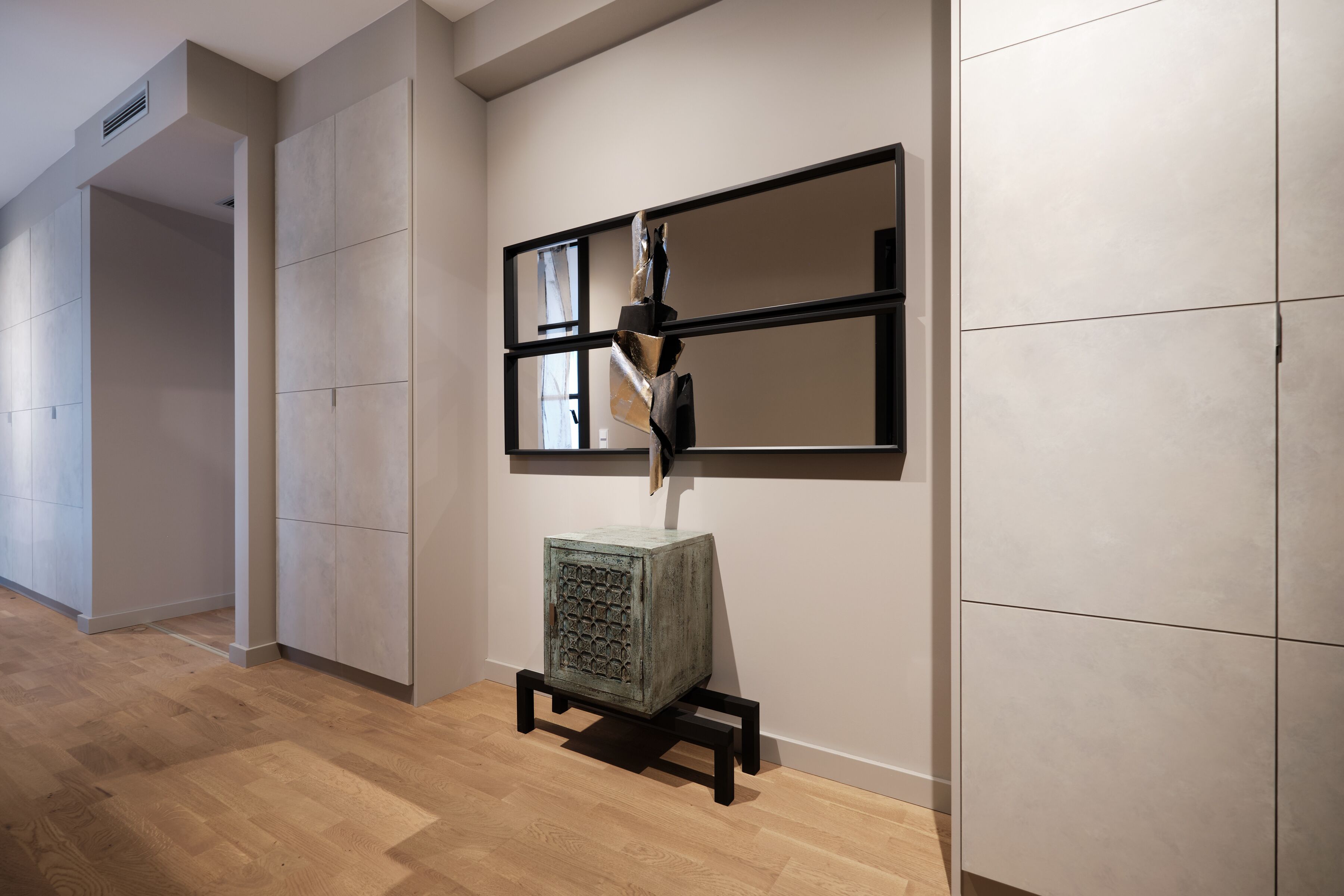 Ein Korridor mit Holzboden, einem grünen Schrank mit strukturierter Oberfläche und einer schwarz-weißen abstrakten Skulptur.