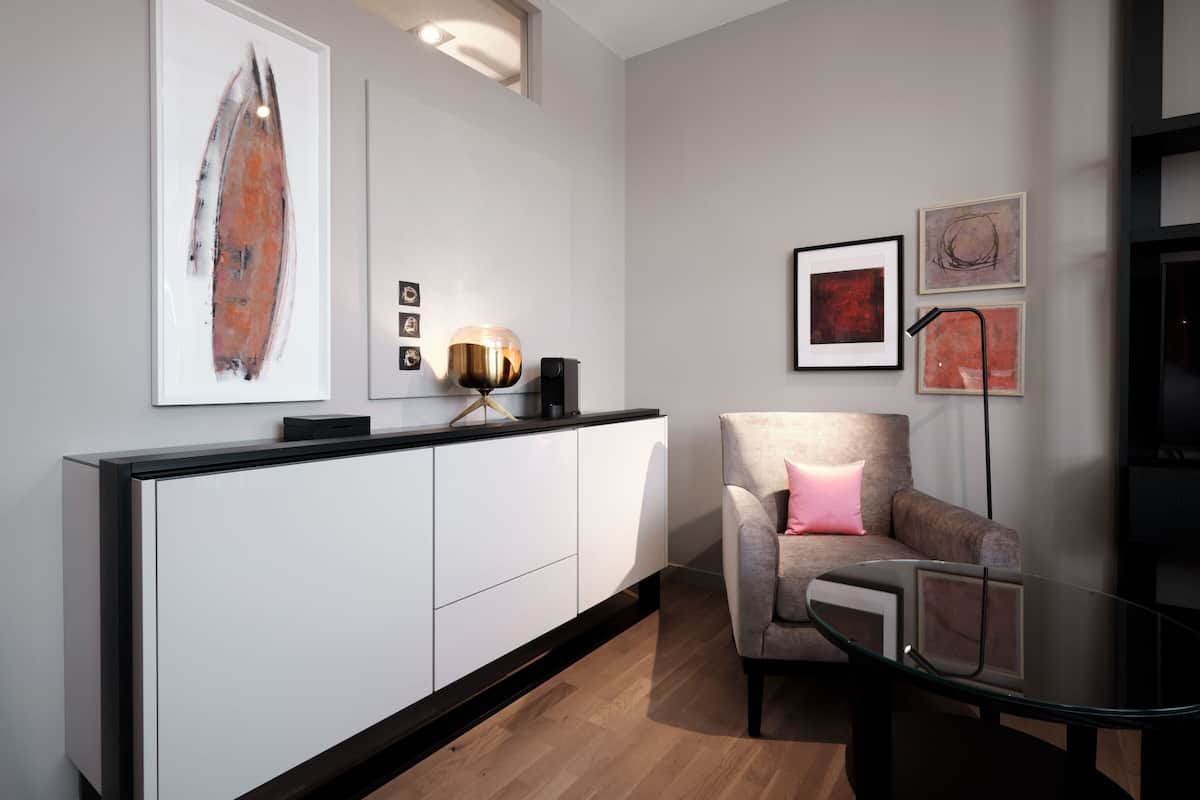 Un salon moderne avec un meuble blanc, une table basse en verre, un fauteuil gris agrémenté d’un coussin rose et des œuvres d’art encadrées accrochées au mur.