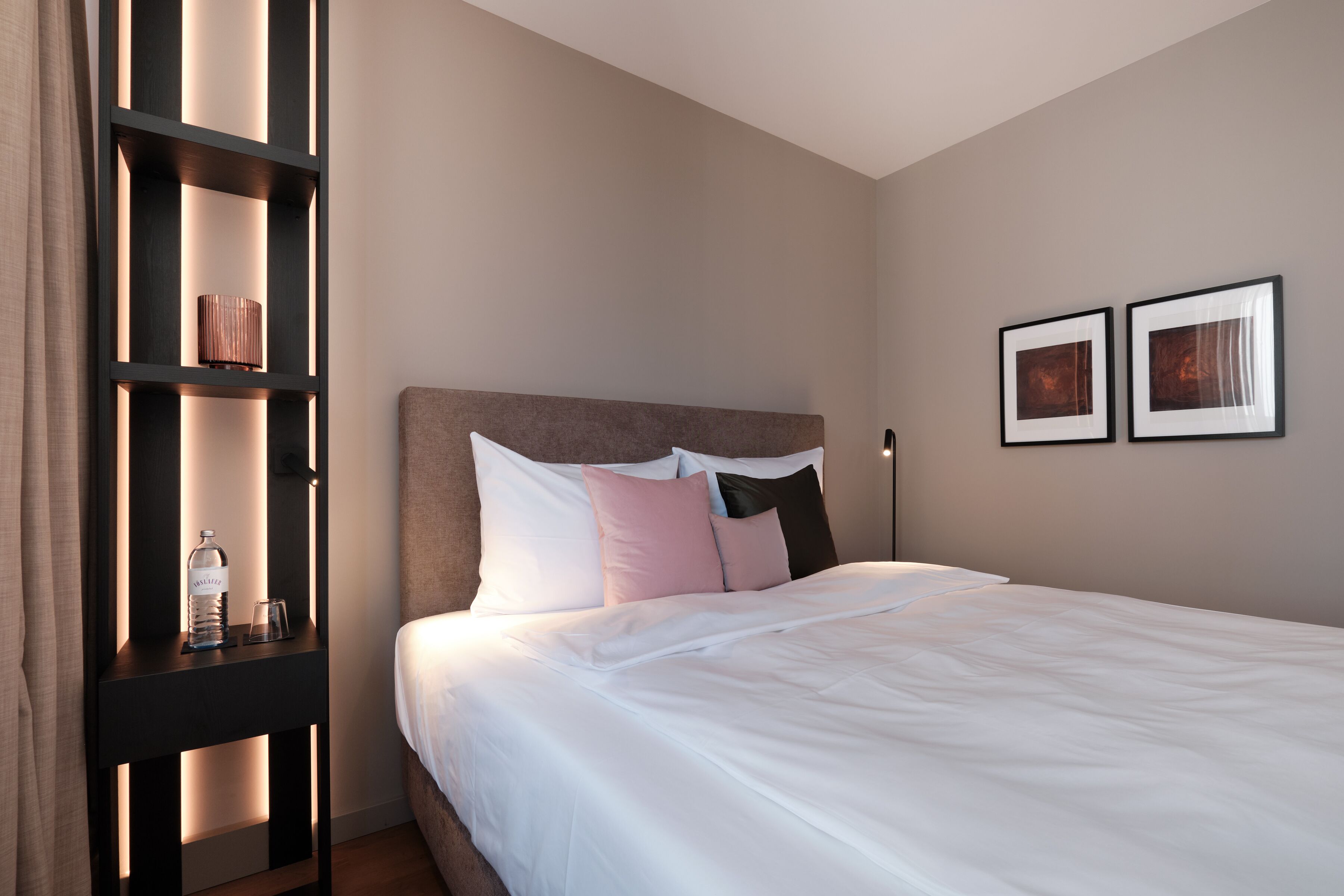 A neatly made bed with white bedding and a headboard, a bedside table with a bottle and glasses, and two framed pictures on the wall.