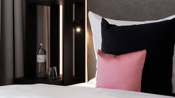 A neatly made bed with a white comforter and two pillows, a dark headboard, and a tall, narrow shelf unit with a bottle and glasses on it.