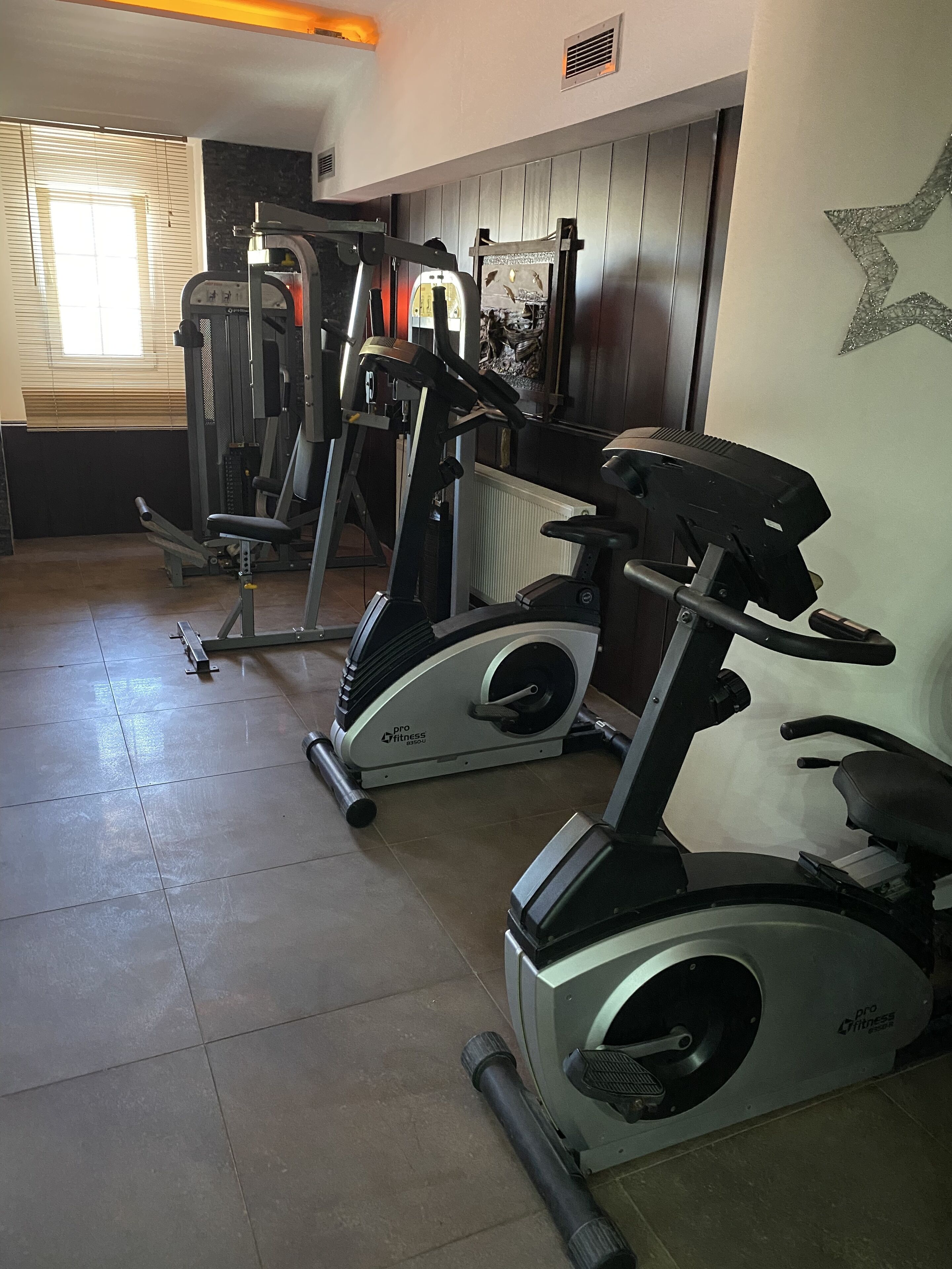 Een fitnessruimte met hometrainers, krachtapparaten en een raam met zonwering.