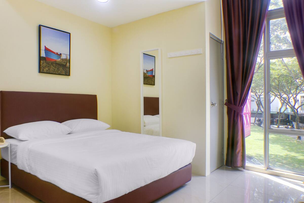 A hotel room with a large bed, a bedside table with a phone, a window with curtains, and a painting on the wall.