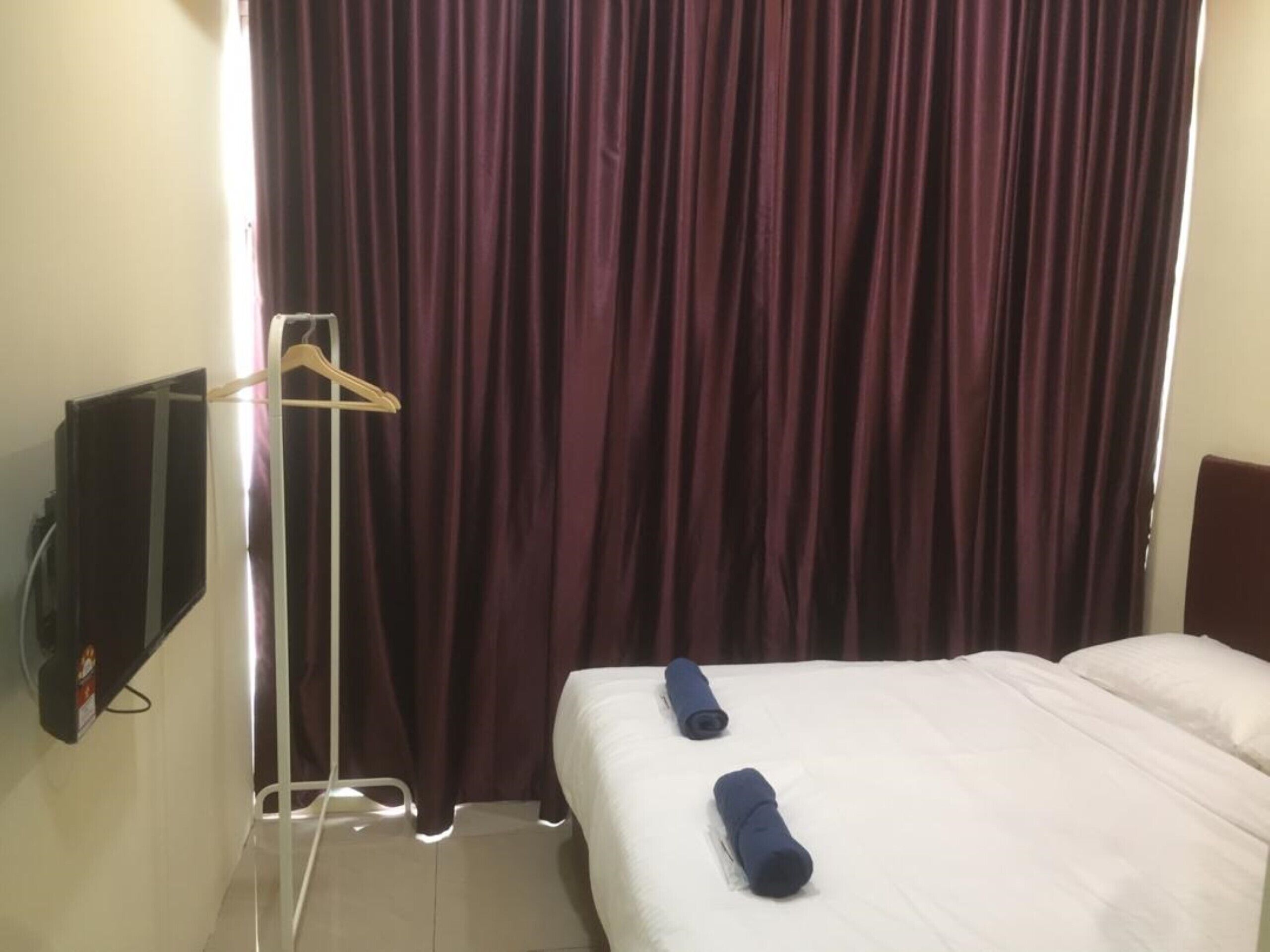 Ein Hotelzimmer mit einem Bett, zwei zusammengerollten Handtüchern, einem an der Wand montierten Fernseher und einem Vorhang.