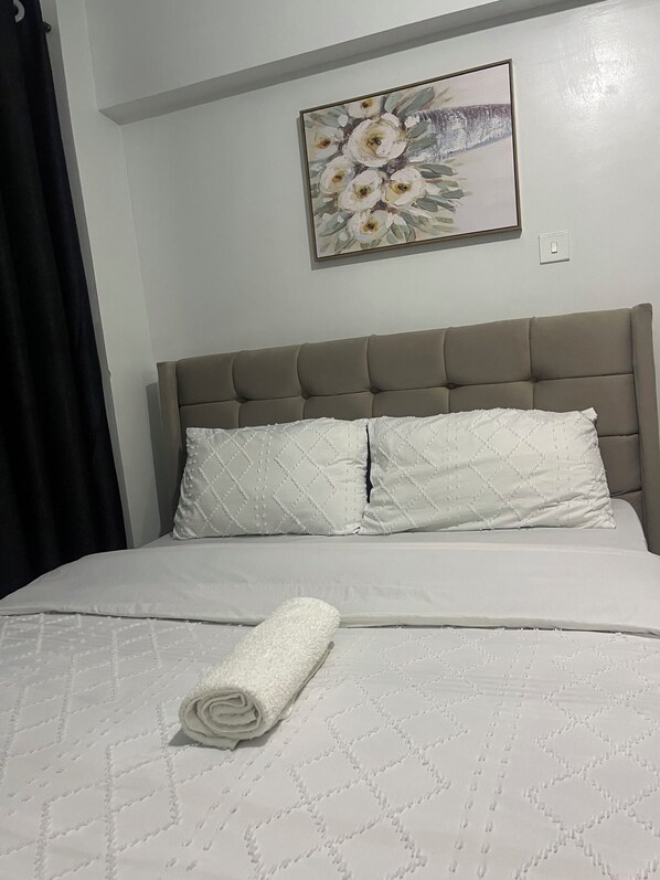 Una cama bien hecha con una toalla blanca enrollada, un cabecero con dos almohadas y un cuadro floral en la pared.