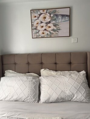 Een slaapkamer met een gebonden hoofdbord, twee kussens met een ruitmotief, en een bloemschilderij aan de muur.