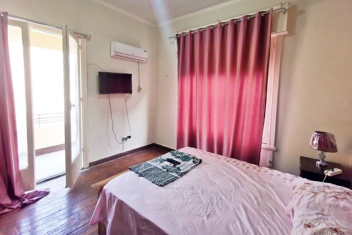 Une chambre à coucher avec un lit, un téléviseur fixé au mur, une fenêtre avec des rideaux et une table de chevet avec une lampe.