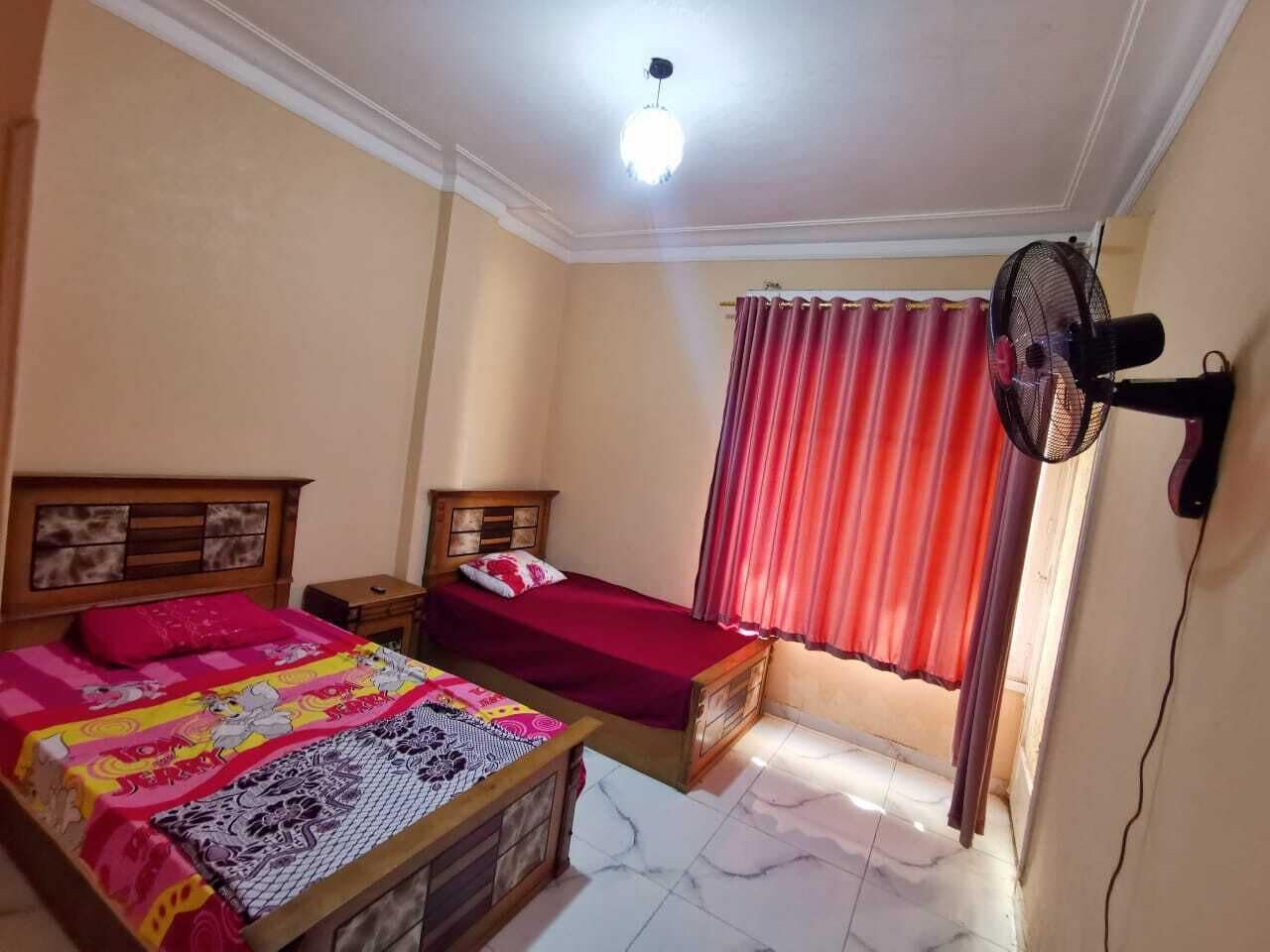 Un dormitorio con dos camas, ventilador de techo y ventana con cortinas rojas.