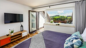 A bedroom with a large bed, a TV, a wooden cabinet, and a window with a view of greenery and buildings. - Oberreute - Modern vacation apartment with panoramic views of the Allgäu (Oberreute)